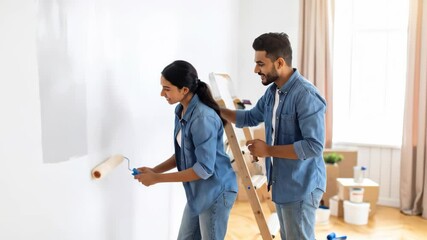 A happy couple paints a white wall together smiling enjoying their home improvement project A ladder and moving boxes sit nearby indicating a recent move - Powered by Adobe