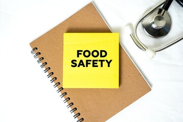 medical context. Shows a yellow sticky note with "Food Safety" written on it placed in a brown spiral notebook, next to a stethoscope.