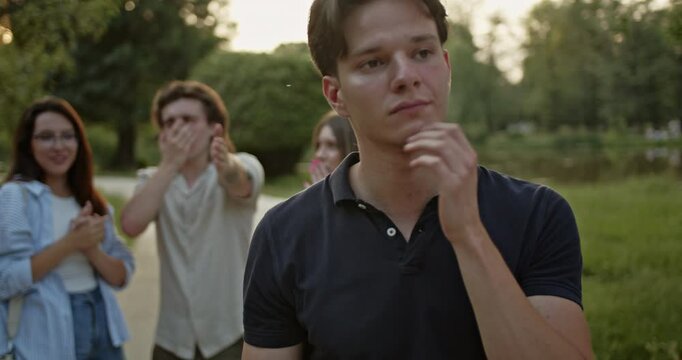 A young man appears deeply sad, wiping his eyes, as his group of friends laugh and tease him in the background. Captures difficult emotions, social dynamics, and peer pressure among young adults.