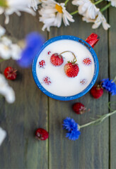 Wild strawberries floating in a blue enamel mug full of milk, placed on a wooden table decorated with daisies and cornflowers, creating a rustic and idyllic breakfast scene