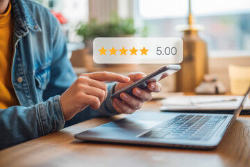 A person uses a smartphone near a laptop, giving a 5-star rating review feedback. Bright, modern workspace suggests online reviews and digital feedback. Ideal for tech or review-focused themes.