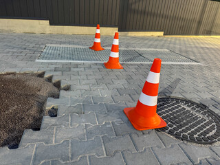 Orange traffic cones mark off a section of uneven pavement near a grate. The bright cones stand out against the gray stones on a sunny afternoon in an urban setting
