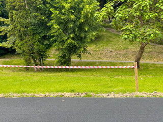 Bright caution tape stretches across a grassy area adjacent to a walking path, signaling restricted...