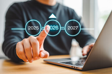 A person interacts with a holographic timeline featuring the years 2025, 2026, and 2027, highlighting 2026. They're using a laptop, symbolizing future technology planning.