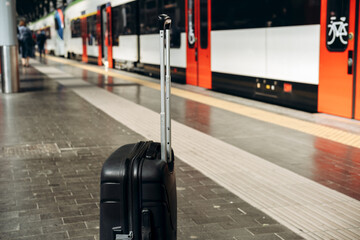 Black rolling suitcase positioned on platform at train station, with blurred train in background,...