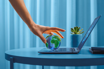 A hand places a miniature green Earth on a laptop keyboard, symbolizing digital connectivity and sustainability. A small plant in the background adds to the eco-friendly theme in the blue setting.