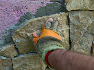 A hand in a work glove lays decorative stone on a wall using cement mortar. The process of...