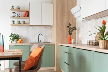 Modern kitchen and wooden dining table with chairs. Green furniture, beige tiles on the wall and orange decoration. New home with elegant design. Real estate.