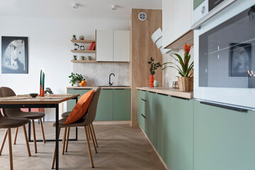 Modern kitchen and wooden dining table with chairs. Green furniture, beige tiles on the wall white oven and orange decoration. New home with elegant design. Real estate.
