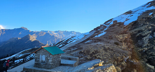 A scenic mountain landscape featuring a stone hut with a green roof nestled along a snowy trekking...