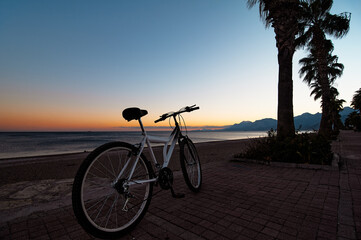 Bicycle parked on a paved path along a beach, silhouetted against a colorful sunset, with palm...