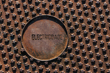 Rusty metal cover on the street, for passing electrical cables with a pattern of stars in relief and the word electricity engraved in Portuguese.