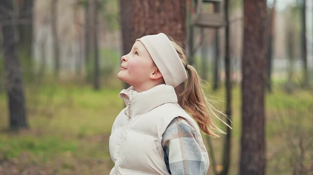 Slow motion view of cute smiling Caucasian schoolgirl walking in the park among trees at autumn day and looking up in the sky. Female kid in jacket and bandage is going near plants and enjoying nature
