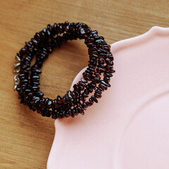 Handmade dark red garnet chip bracelet coiled on soft pink ceramic plate, wooden background. Concepts: fashion accessory, gift idea, handmade jewelry, product display.