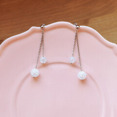 Two silver chain earrings with clear crackle glass beads placed on elegant pink ceramic plate. Warm wooden background. Concept of handmade jewelry, product styling, modern elegance, soft aesthetic.
