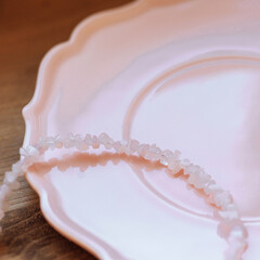 Light pink crystal chip necklace on a pastel pink ceramic plate over wooden floor. Natural stones, soft tones. Jewelry concept, handmade style, boho aesthetics, wellness. With copy space.