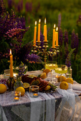 Romantic table decor for a loving couple on the blooming meadow with purple lupines. Two glasses of limoncello, flowers in a vase, candles, lemons, wooden furniture. Sunset, golden hour.