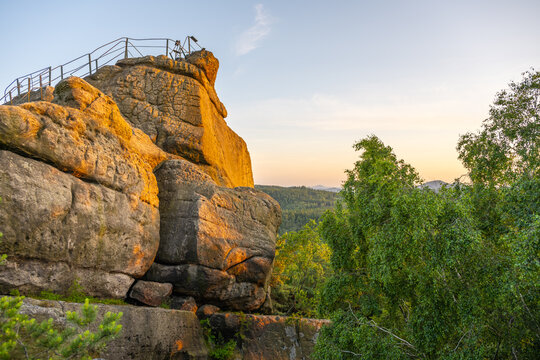 The warm glow of sunset illuminates the sandstone Popov Cliffs in Czechia. Lush greenery surrounds the rocky formation, showcasing the beauty of nature in the evening light.