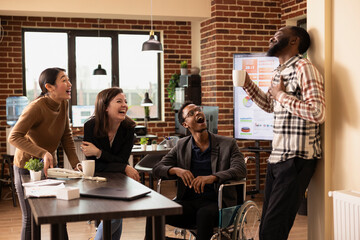 Multiracial group of professionals in brick wall office take a break, chatting and laughing together. African American man with coffee cup shares an amusing story with his team.