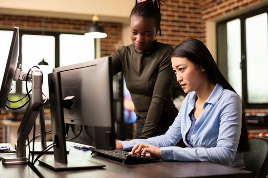 Diverse women collaborating at computer, engaged in a creative discussion in modern office. Closeup of asian employee typing as african american manager observes and reviews work on a desktop monitor.