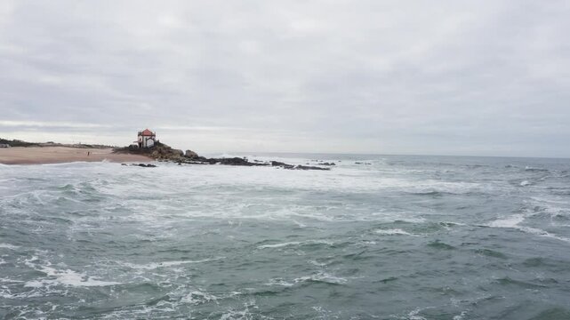 Capela do Senhor da Pedra church on the sandy pinissula washed by winter cold Atlantic ocean waves on Praia do Senhor da Pedra in Nothern Portugal. Traveling and beauty in Nature concept slow drone 4K