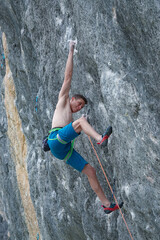 CLOSE UP, LOW ANGLE VIEW: Shirtless climber extends his arm, reaching for a distant hold on a steep...