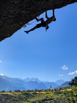 LOW ANGLE VIEW, SILHOUETTE: Inspiring alpine scenery with mountain peaks and charming village beneath a rock climber hanging from an overhang. Stunning beauty of popular sport climbing crag in summer