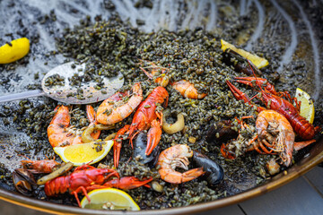 Black seafood paella with shrimp, squid, mussels, lemon wedges served in large pan at food event. Concept of black paella, marine flavors, festive culinary presentation