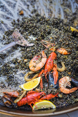 Close-up of black seafood paella with shrimp, crayfish, lemon slices, squid served from large metal pan. Concept of seafood paella, rich flavor, Mediterranean cuisine tradition