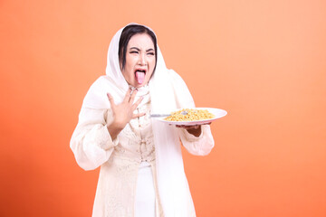 A woman wearing hijab looks disgusted, holding a plate of noodles, making a face, sticking her...
