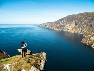 Teenager girl sightseeing stunning Slieve League cliff nature scenery and Atlantic ocean. Blue cloudy sky. County Donegal, Ireland. Unique landmark and tourist point of view and hiking area.