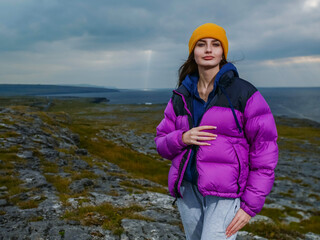 A girl is wearing a purple jacket and a yellow hat stands on a rocky hill overlooking the ocean. The scene is serene and peaceful, with the woman posing for a photo. Burren area, Ireland. Travel 