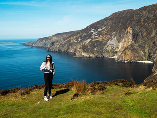 Teenager girl sightseeing stunning Slieve League cliff nature scenery and Atlantic ocean. Blue cloudy sky. County Donegal, Ireland. Unique landmark and tourist point of view and hiking area.