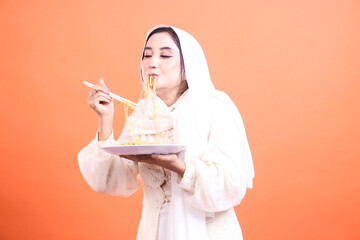 A beautiful muslim woman in hijab eating a plate of noodles using chopsticks in studio. isolated on a orange background.