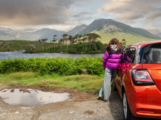 Young teenager girl standing by small red sport car and enjoys view of Twelve pines island and mountains in Connemara, county Galway, Ireland. Famous tourist area with amazing nature scenery.