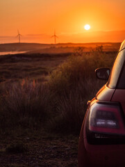 A red car is parked in a field with wind turbines and a warm sunrise in the background. The car is facing the sun, and the sky is orange and pink. The scene is peaceful and serene. Early bird concept