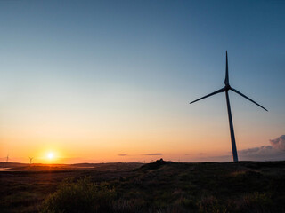 Silhouette of a tall wind turbine against blue sky and warm rising sun. Renewable source of energy...
