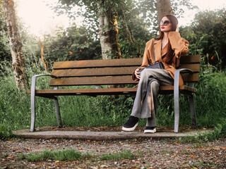 Young and stylish teenager girl sitting on a bench in a park alone. Waiting for date concept. The model wears trendy brown jacket and dark glasses and looks calm and elegant. Selective focus.