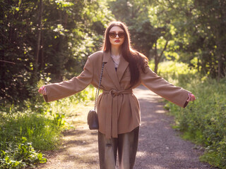 Young teenager girl in trendy brown jacket and black handbag and hands up is standing on a footpath in a park with warm sunny glow in the background. Fashion and beauty. Model enjoy fine summer day.
