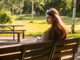 Stunning young teenager girl is sitting on a bench in a park with her smart phone in her hand, warm glowing summer background. Selective focus. Model looks calm and thoughtful. Enjoy shade in heat