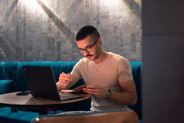 Focused young man with glasses writing in a notebook while working on a laptop in a modern cafe....