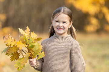Happy smiling little girl in a cozy brown sweater holds a bouquet of vibrant yellow autumn leaves...