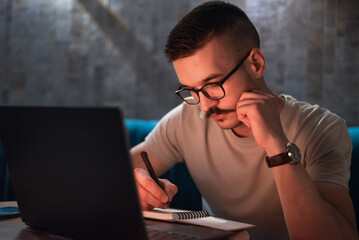 A young man wearing glasses writing in a notebook while working late on a laptop or a male student...