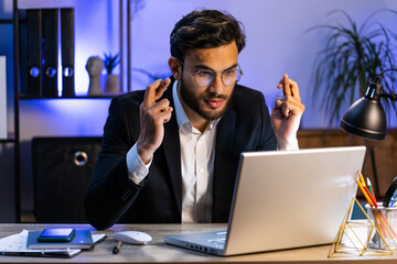 Excited Indian businessman crossed fingers looking at laptop screen asking for good luck news,...