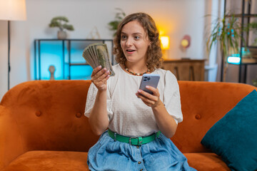 Successful rich woman holding smartphone and waving dollar bill money fan sitting on sofa couch at...
