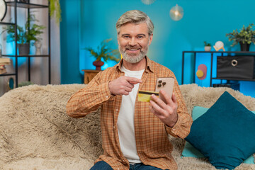 Caucasian mature man sitting on home sofa, holding smartphone and credit card, making money...