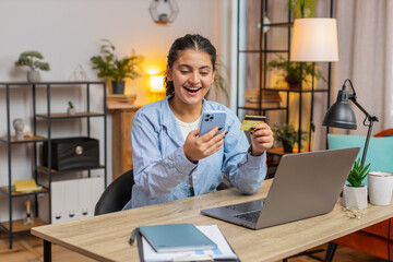 Happy Indian woman freelancer using bank credit card and smartphone for online shopping payments at...