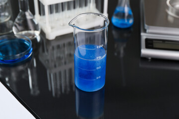 Beaker with blue liquid, other laboratory glassware and scale on black mirror table, closeup