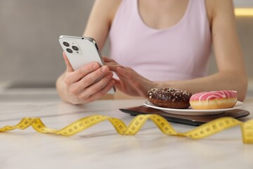 Woman using smartphone at white marble table with kitchen scale, donuts and measuring tape indoors,...