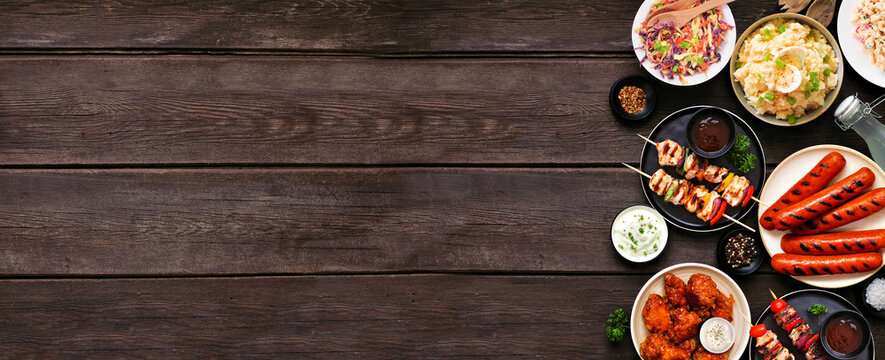 Summer BBQ food side border over a dark wood banner background. Assorted grilled meats, salads and sides. Top view.